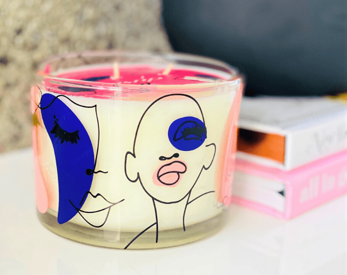 A candle in a glass jar features abstract face art with bold blue and pink shapes. Two stacked books are partially visible in the background.