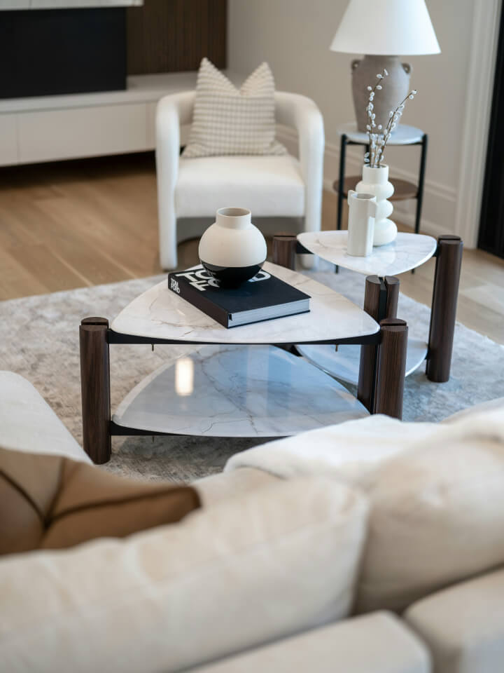 Modern living room with a marble coffee table, decorative vases, a stacked book, a white armchair, and a beige sofa on a light rug.