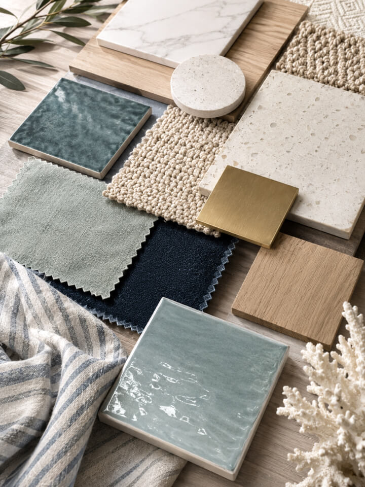A flat lay of various interior design samples, including fabric swatches, tiles, wood, carpet, and metal finishes in neutral, blue, and green tones arranged neatly on a surface.