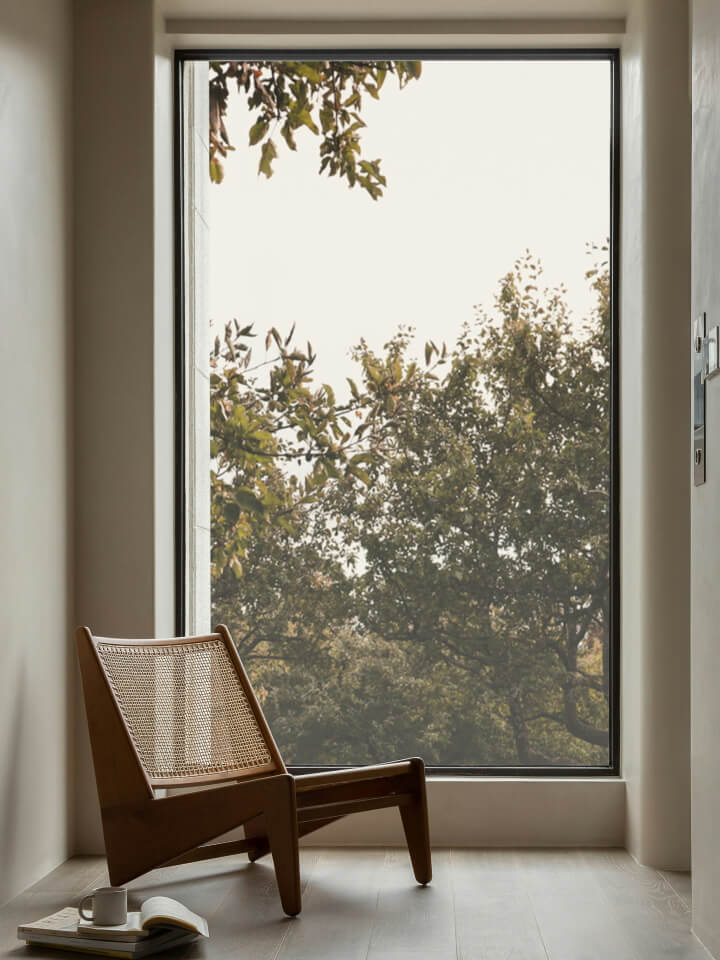 A wooden chair sits on a light floor near a large window overlooking leafy trees. An open book and mug rest beside the chair.