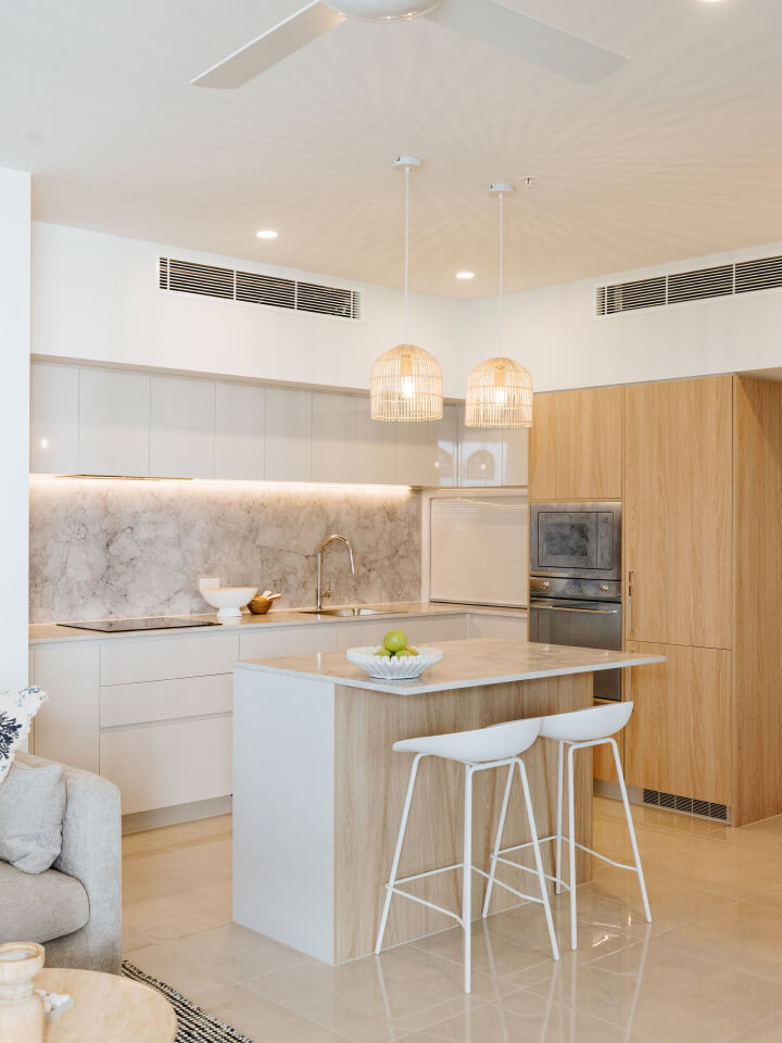 Modern kitchen with wood and white cabinetry, marble splashback, built-in oven, island with two white stools, and pendant lights overhead.
