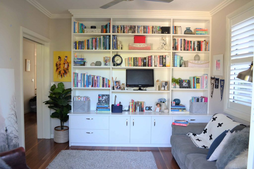 Living room with white built-in shelves filled with books, decor, a small TV, cupboards below, a grey sofa, a potted plant, and a window with white blinds.