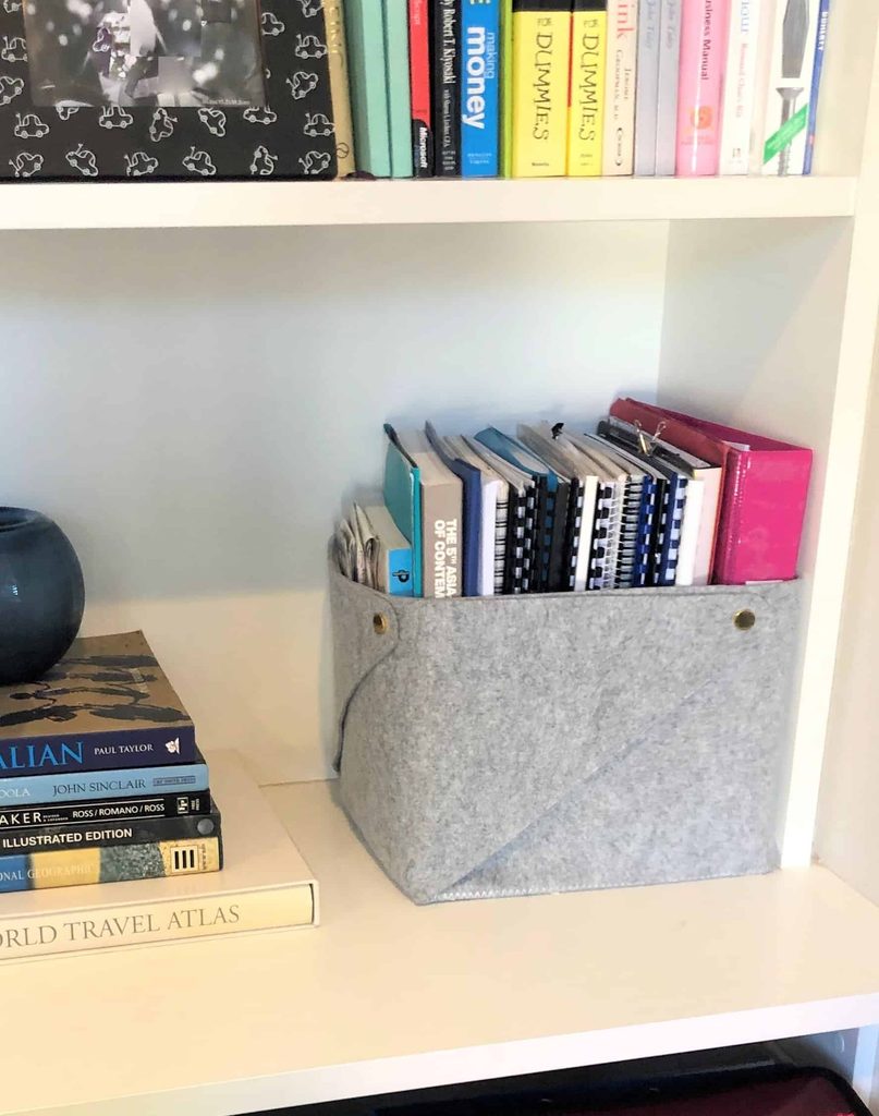 Grey felt organiser on a white shelf holds notebooks and folders; nearby are stacked books, a blue vase, and an atlas.