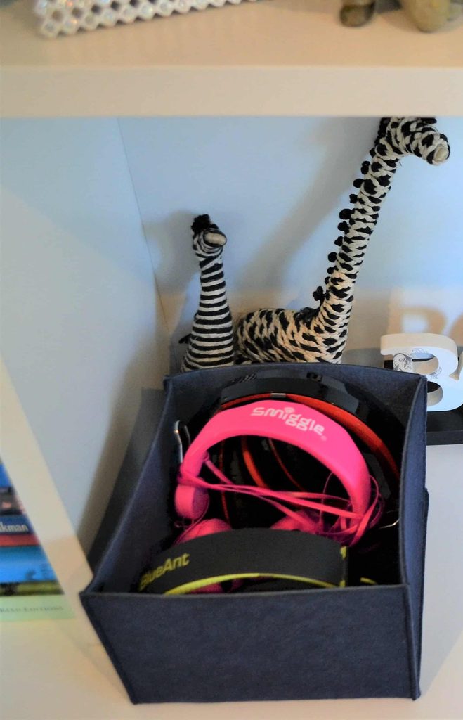 Two toy giraffes stand behind a fabric box containing colourful headphones on a white shelf.