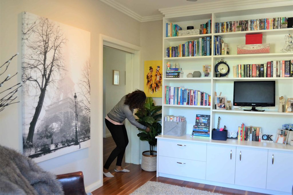 A person adjusts a potted plant next to a built-in bookcase filled with books and decor in a well-lit living room.