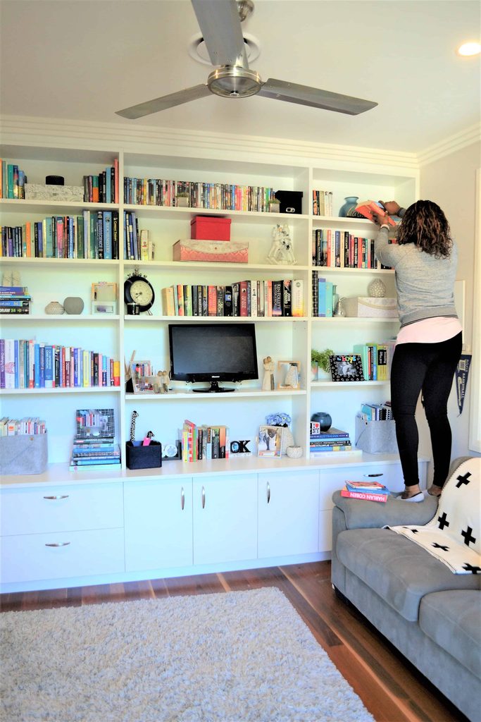 A woman stands on a sofa whilst organising items on the top shelf of a large built-in bookcase in a lounge.
