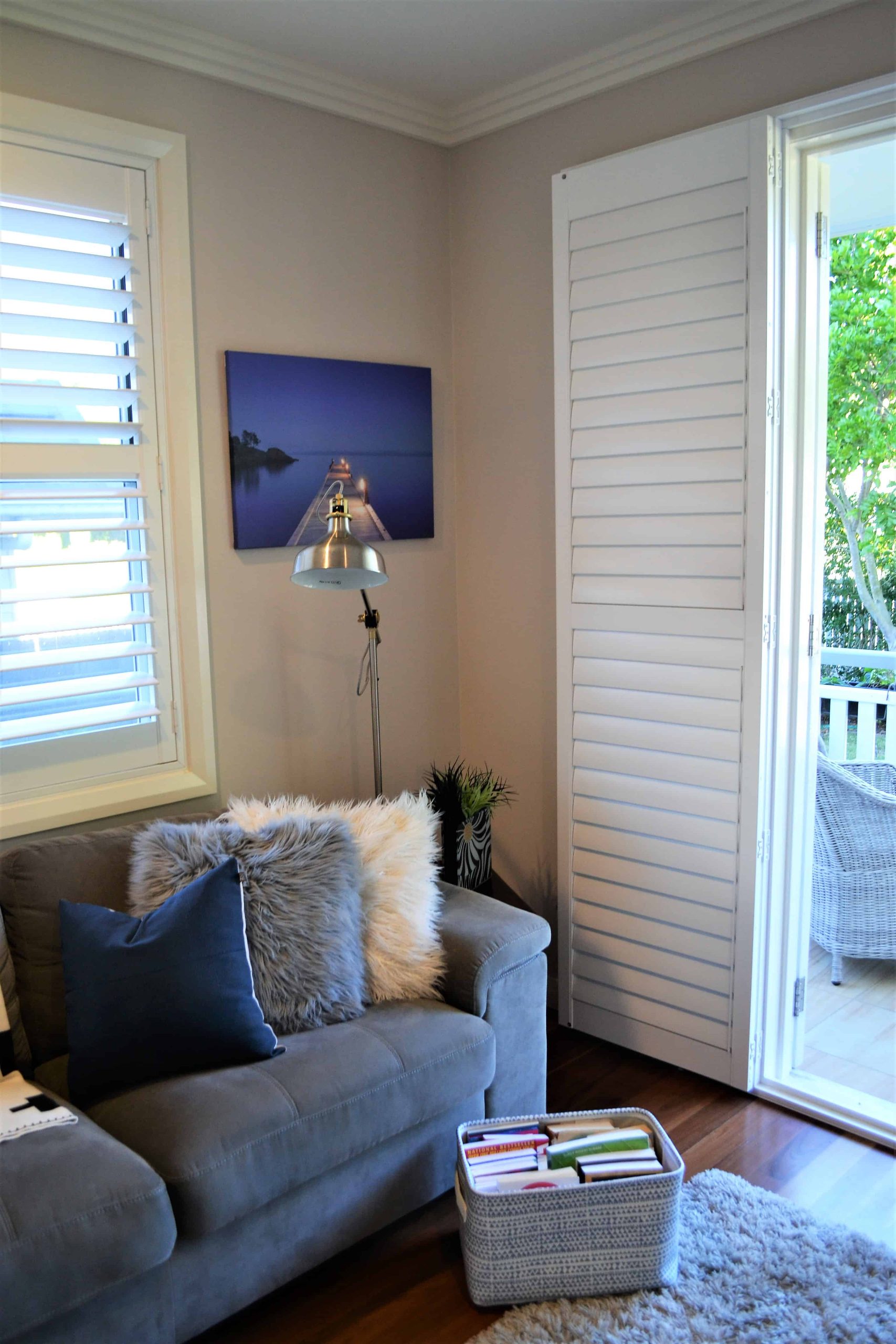 A cosy living room corner with a grey sofa, fluffy cushions, a floor lamp, wall art, and a window with white shutters open to a porch and greenery outside. A basket of books sits on the floor.