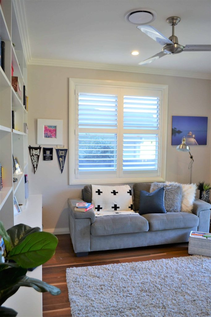 A grey sofa with black and white cushions sits by a window with white shutters; a floor lamp and bookcase are also visible in a cosy, well-lit living room.