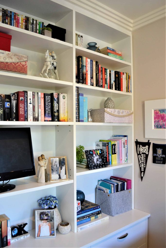 White bookcase filled with books, storage boxes, decorative items, a framed photo, a figurine, a small telly, and some flowers, next to a wall with a framed picture and pennants.