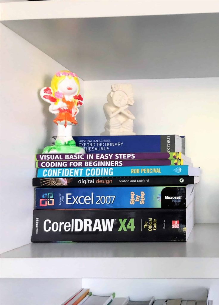 A stack of books on coding, design, Excel, and a dictionary sits on a shelf with one clay and one marble doll on top.