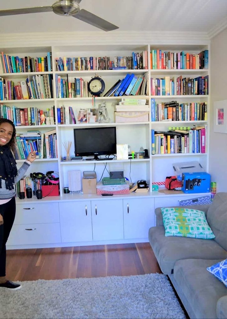 A woman stands smiling in front of a large white bookcase filled with books and various items, next to a grey sofa with colourful cushions in a cosy, well-lit room.