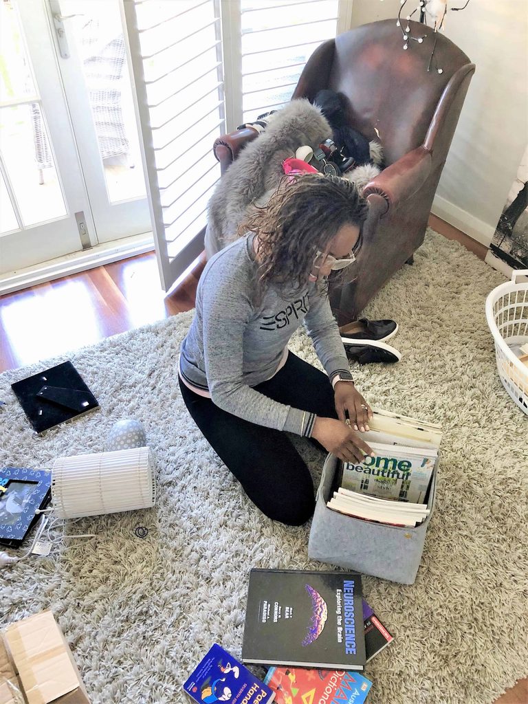 A person kneels on a carpet, sorting magazines in a bin. Around them are books, laundry baskets, and household items in a well-lit lounge.