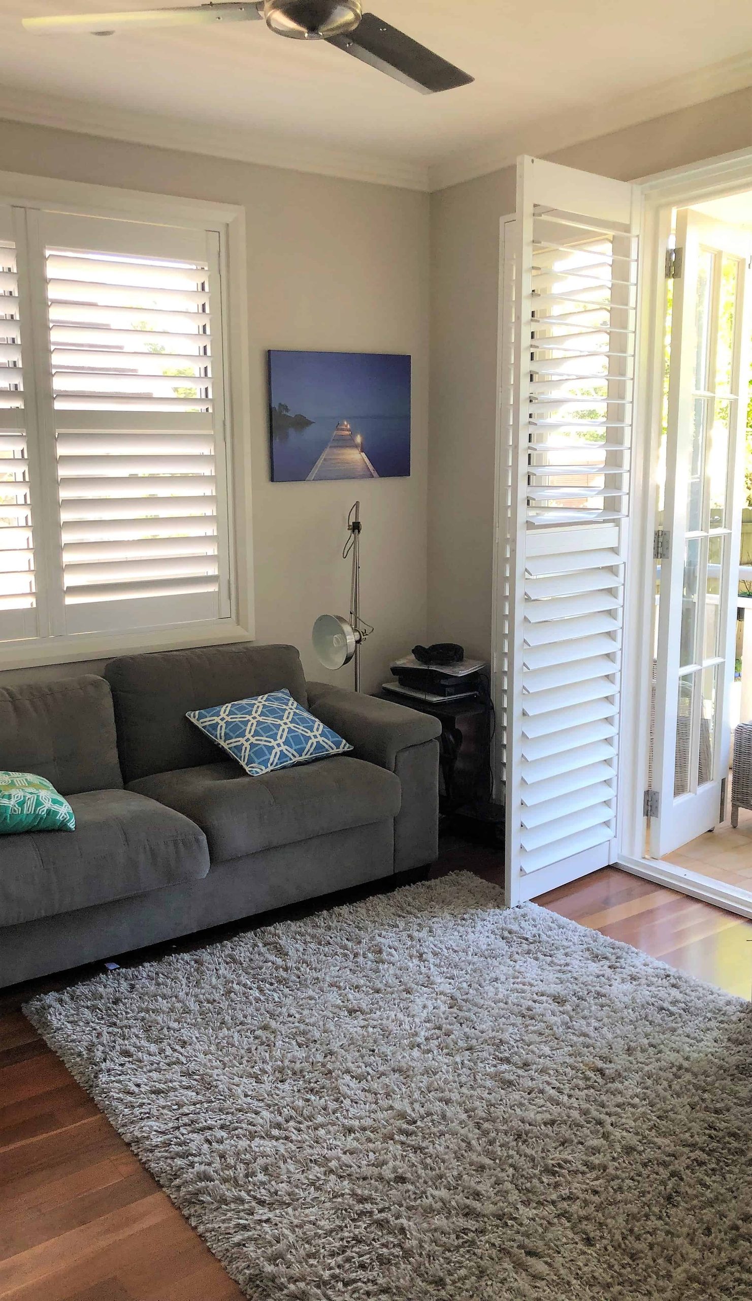 A living room with a grey sofa, two patterned cushions, a floor lamp, and a large grey rug, with sunlight coming through open shutter doors.