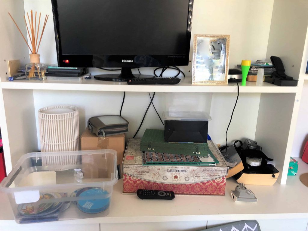 A desk with a telly, framed photo, remote, plastic containers, a lamp, and various office supplies arranged on shelves.