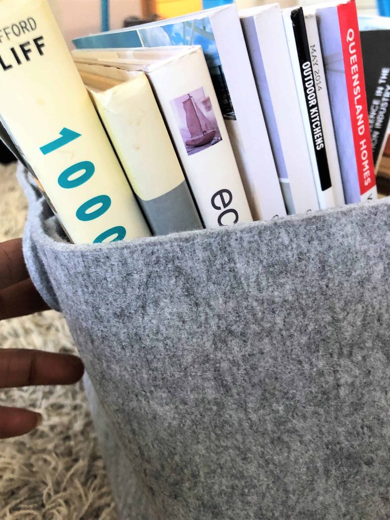 A grey felt basket filled with upright books and magazines, with a person’s hand holding the edge on the left side.