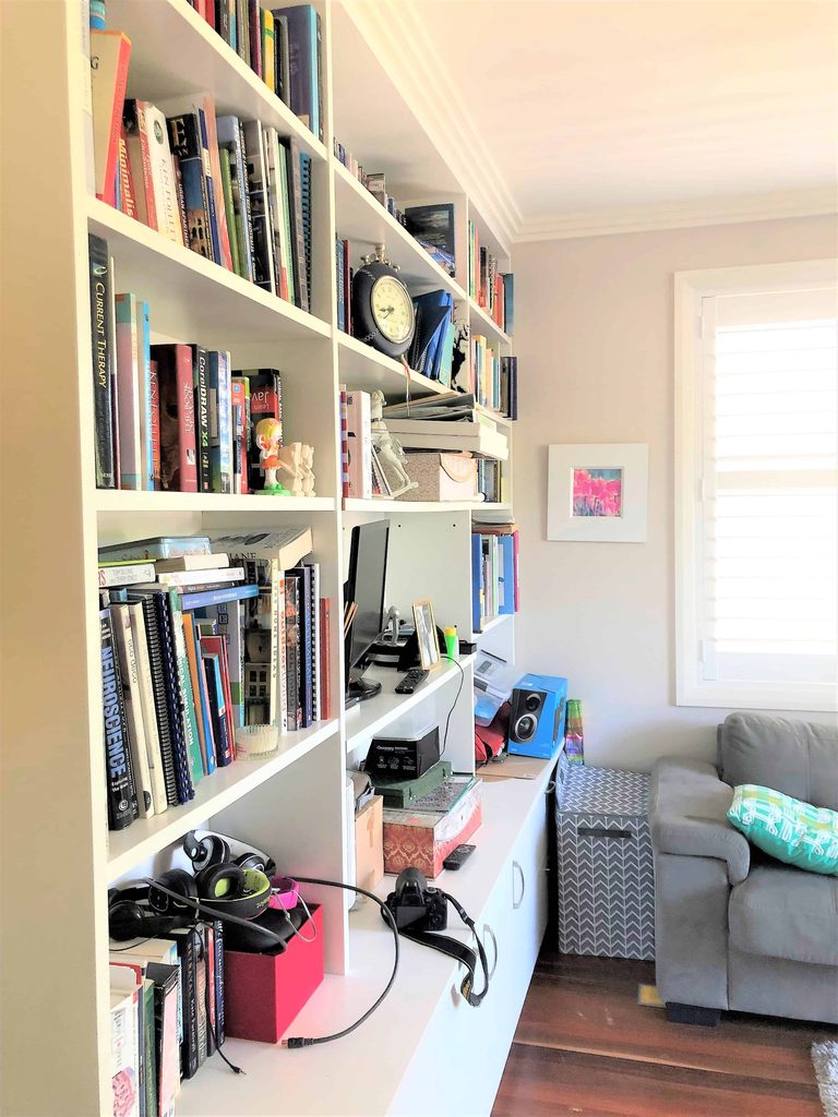 White shelves filled with books, a clock, and other items. Below, a cluttered desk with electronics and headphones. A grey sofa and window are on the right.