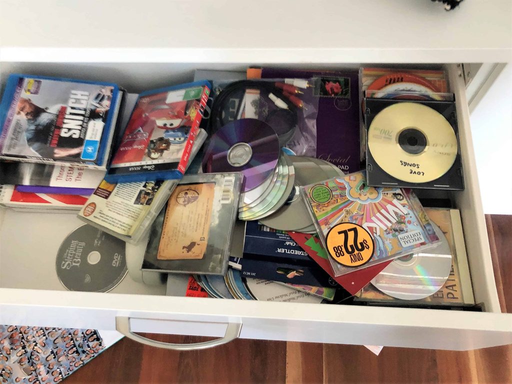 An open drawer filled with assorted CDs, DVDs, cases, and some scattered paper items, organised in a random manner.