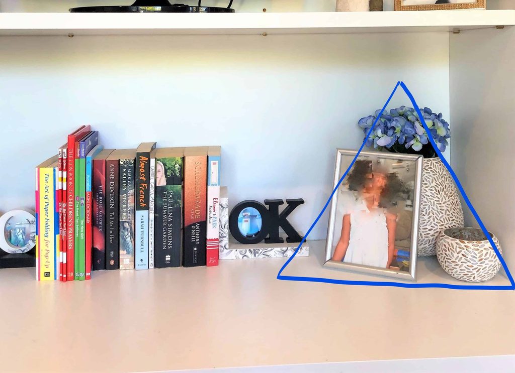 A shelf with a row of books, a framed blurred photo, a small blue flower arrangement, a white decorative vase, an “OK” sign, and a round object.