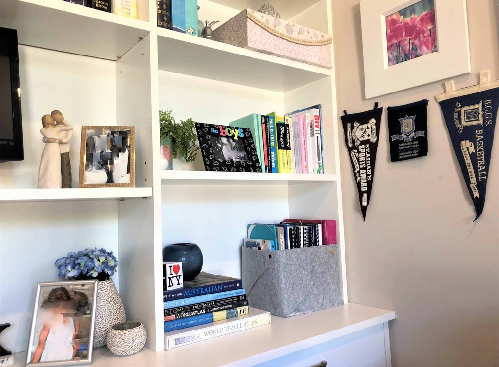 White shelving unit with books, framed photos, decorative objects, a felt storage bin, and pennants on the wall. A desk below holds stacked books, a vase, and a blue artificial flower arrangement.
