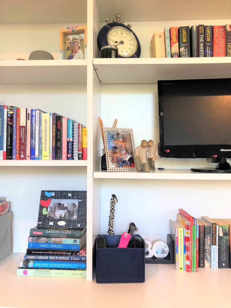 White shelving unit with books, a clock, TV, framed photo, figurines, stacked magazines, a pen holder, a toy giraffe, and small decorative items on a white desk surface.