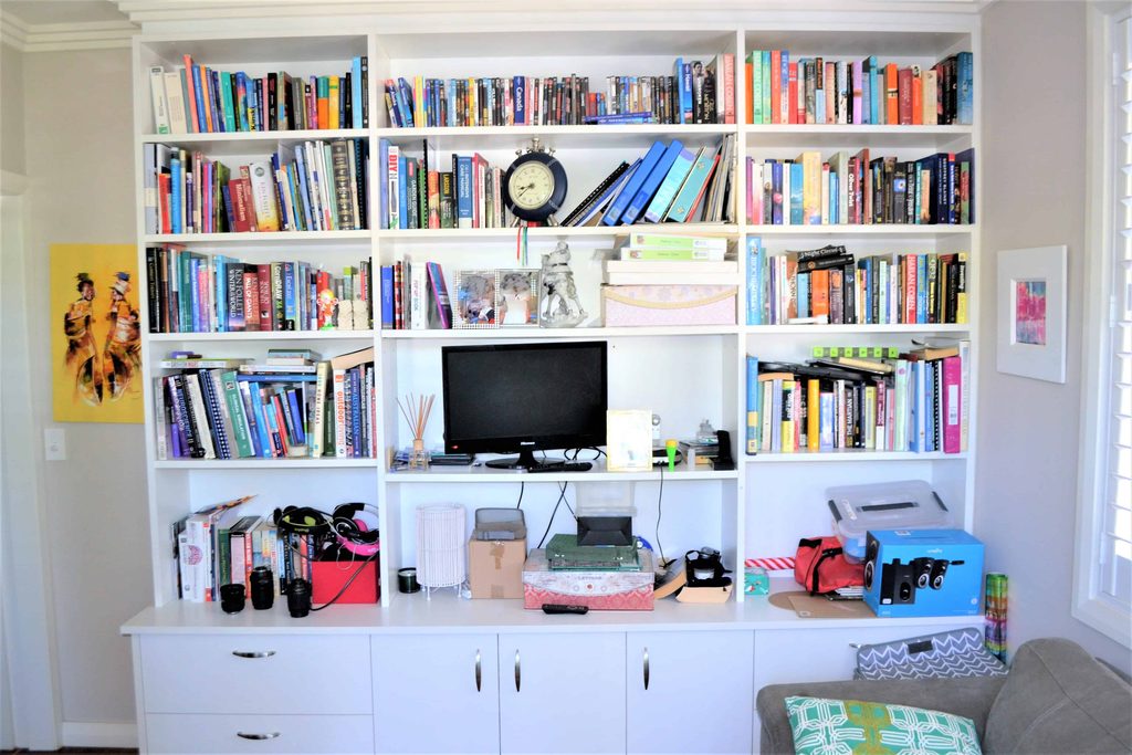 White bookcase filled with books, a clock, computer monitor, office supplies, cameras, and various items on the shelves and worktop.