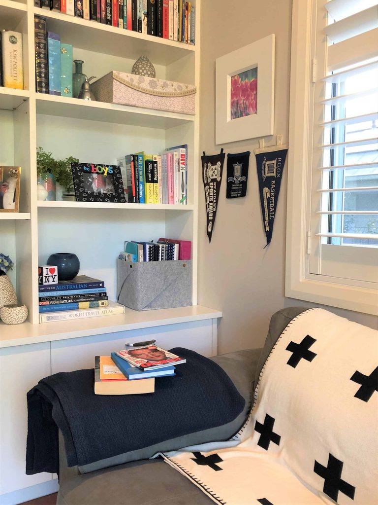 Bookshelves with books and decor items, pennants on the wall, and a sofa with a black-and-white blanket and a stack of books near a window with white shutters.