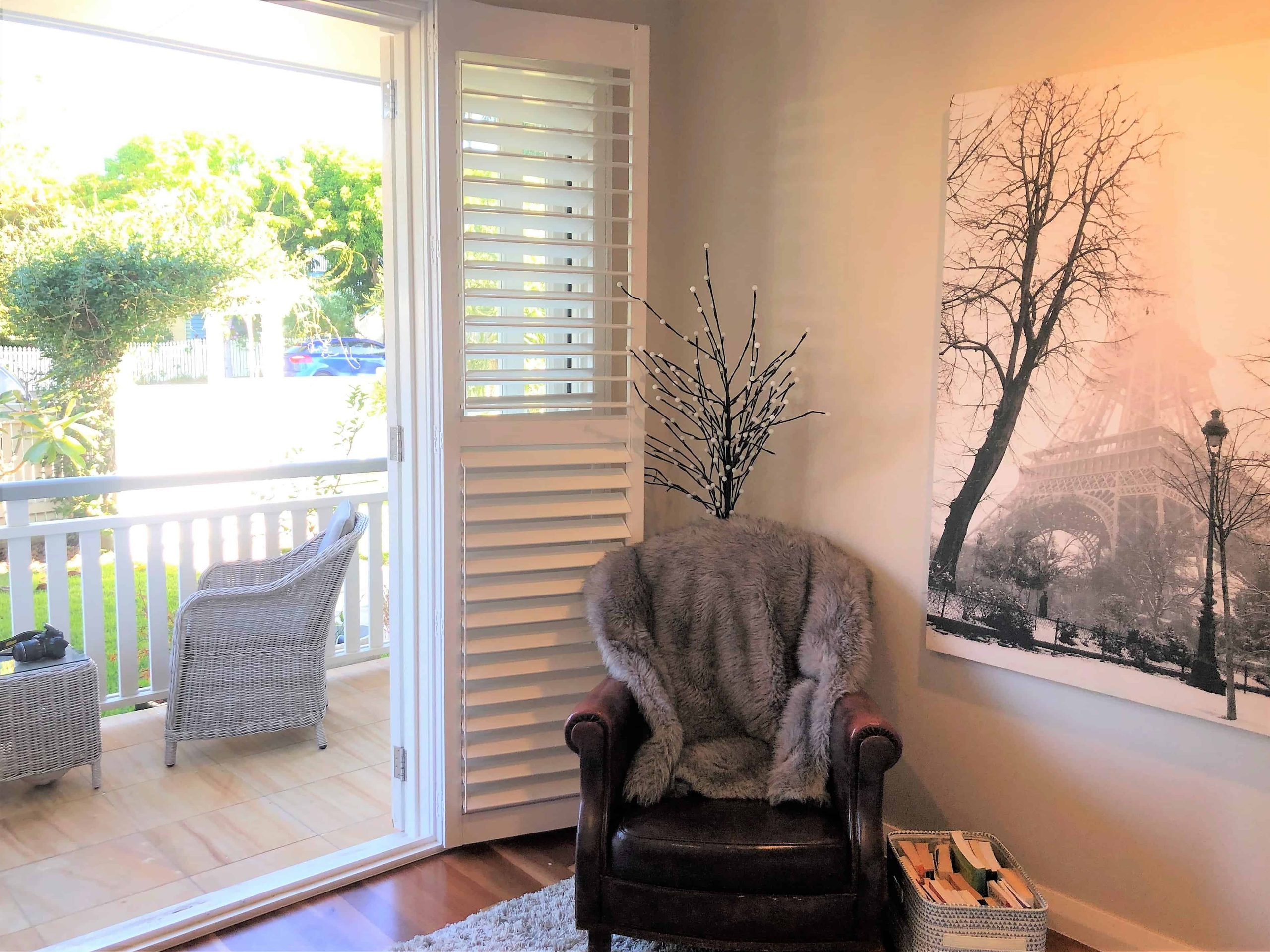 A cosy corner with a leather chair draped in fur, next to a large black-and-white Paris photo, beside open shutters leading to a sunlit porch with wicker furniture.