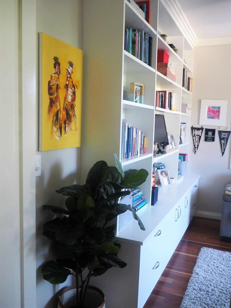 Tall shelves with books and decorative items line a wall; a plant in a basket sits below a colourful painting of two figures on a yellow background.
