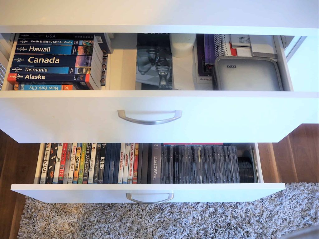 Two open white drawers; the top contains books, notebooks, and a case, while the bottom holds a row of CDs and DVDs.
