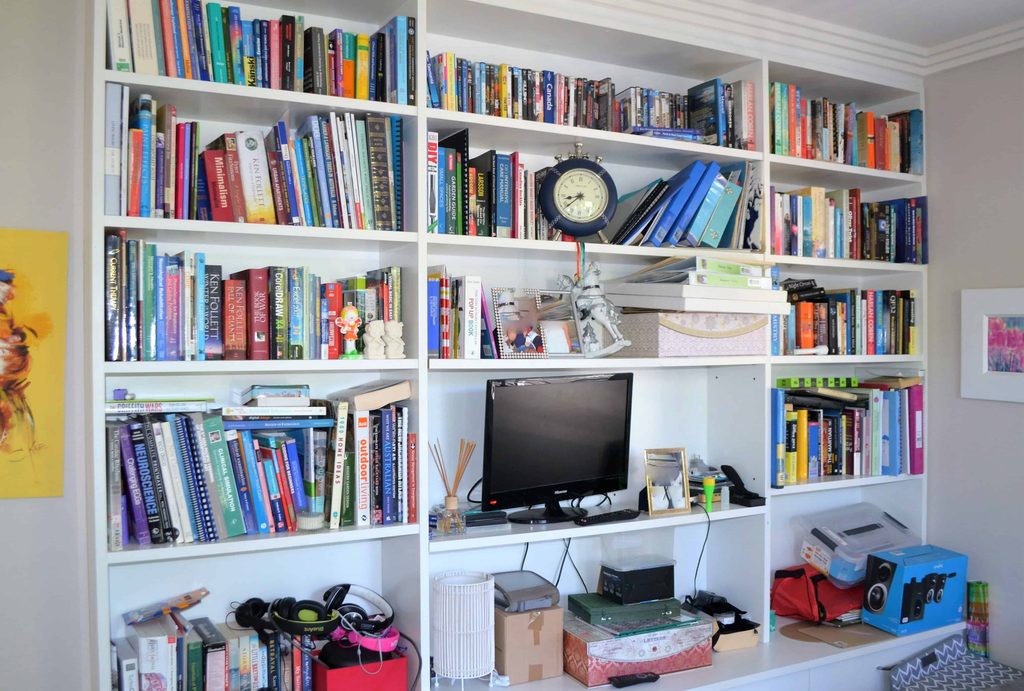 White bookcase filled with colourful books, a clock, a computer monitor, boxes, office items, electronics, and decor items arranged across multiple shelves.