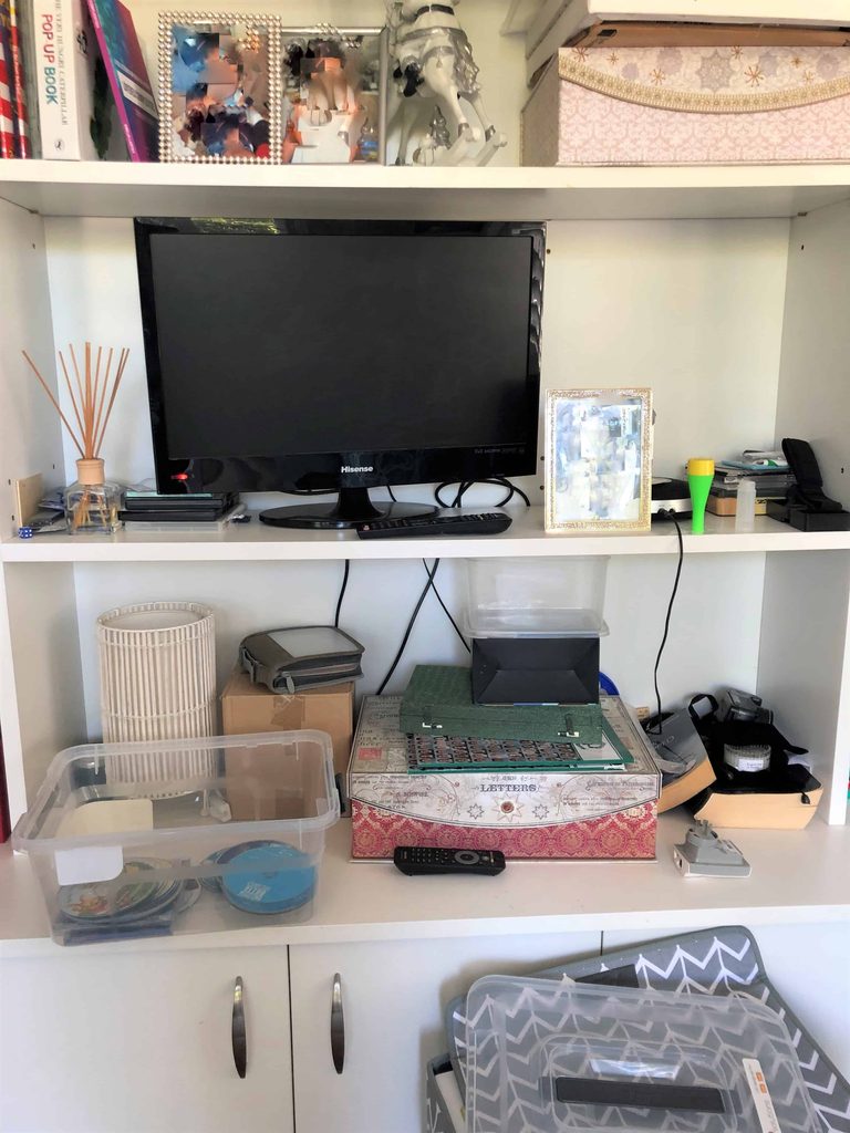A desk shelf with a monitor, framed photos, boxes, organiser trays, electronics, and assorted office supplies arranged on three shelves.