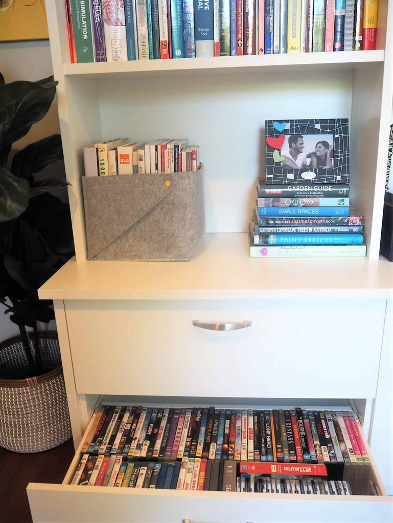 White bookcase with books on top shelves, a felt box with small books, a stack of books, and an open drawer filled with DVDs. A leafy plant is partially visible on the left.