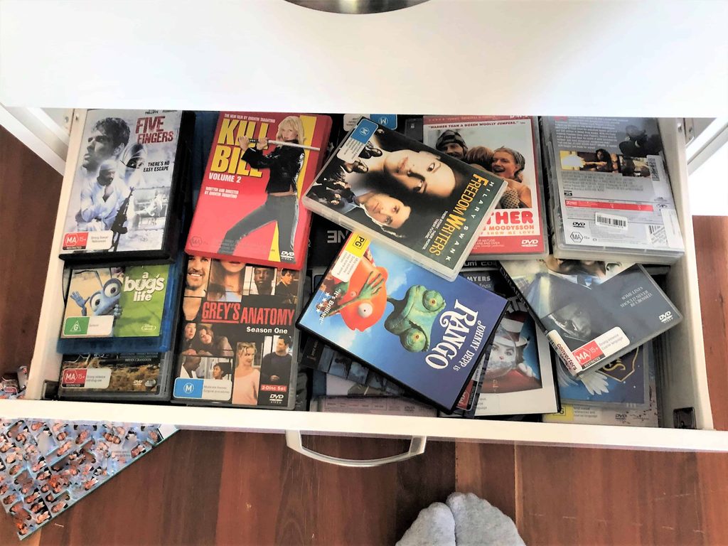 An open drawer filled with assorted DVD cases, including films and TV series, with a pair of feet visible at the bottom edge of the image.