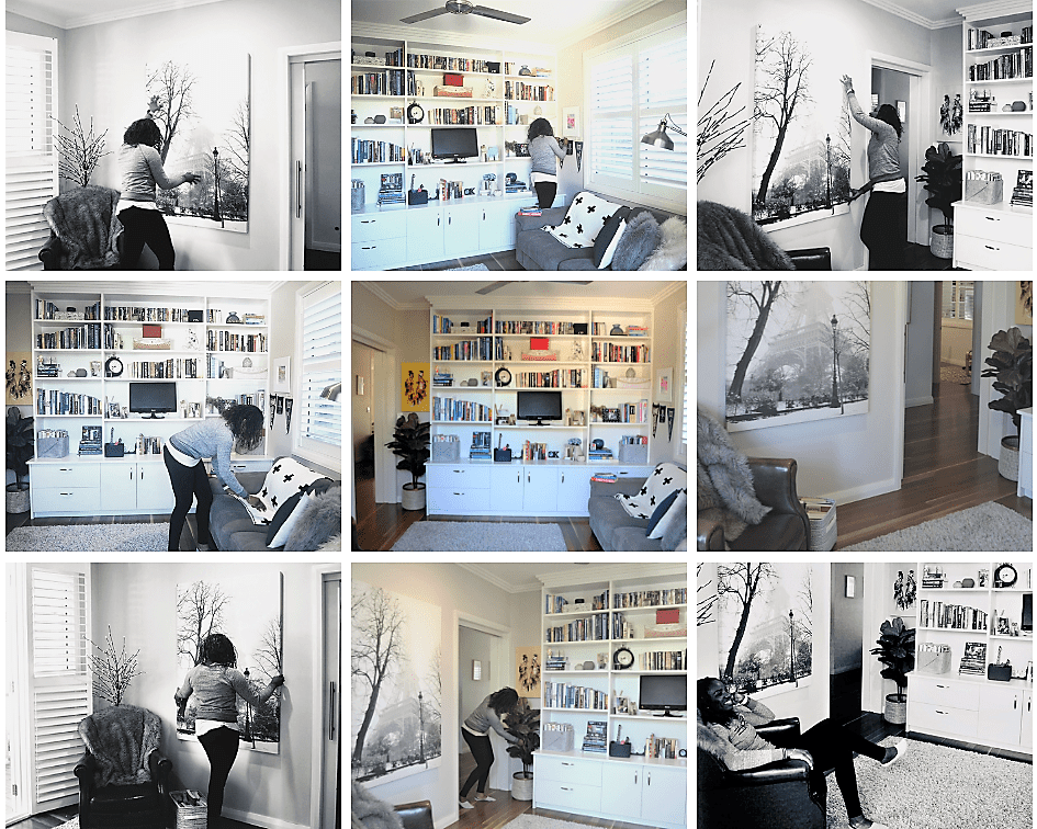 A person arranges art on the wall and shelves in a bright living room with bookcases, large windows, and modern décor.