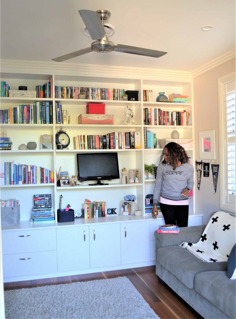 Woman stands next to a built-in bookcase filled with books and decor items in a lounge with a sofa, rug, and ceiling fan.