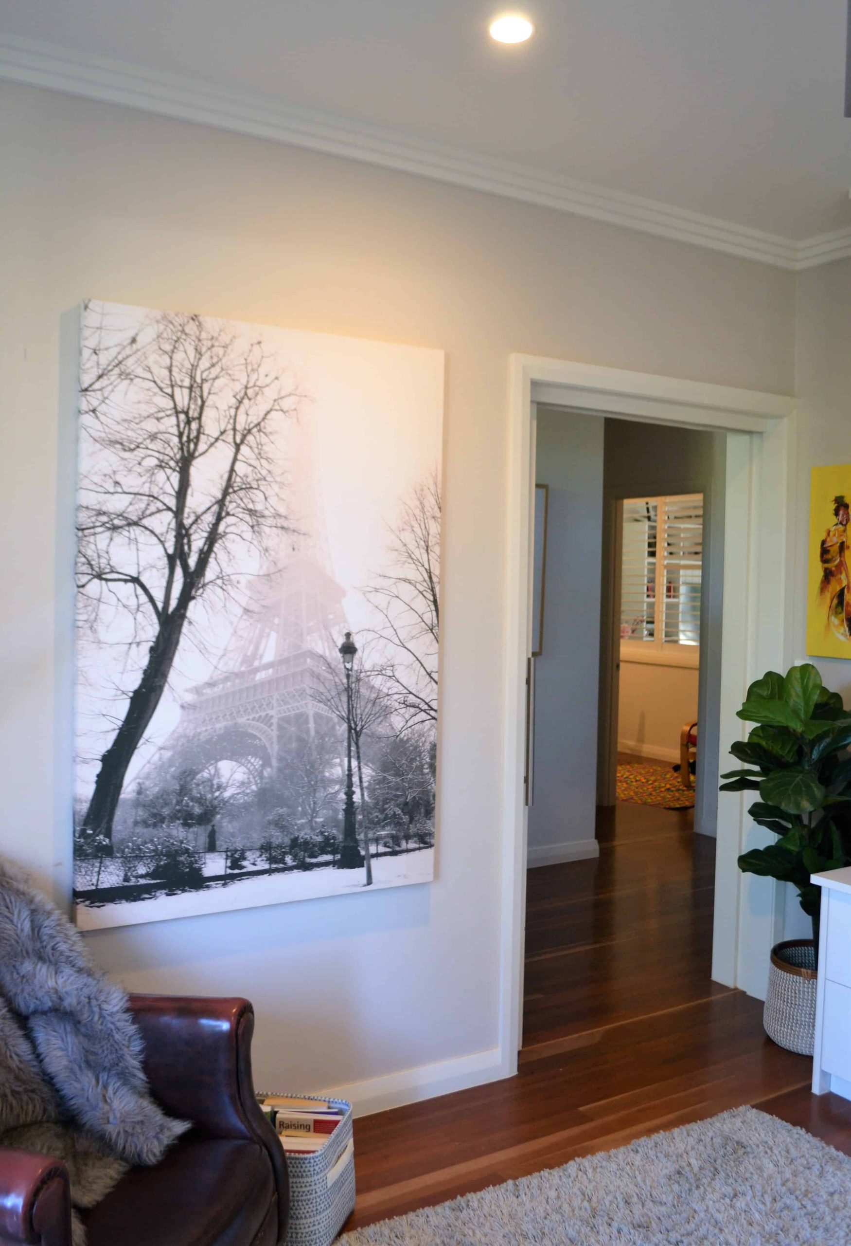 A modern living space with a large black-and-white Eiffel Tower photo on the wall, wooden floors, a plant, and a view into an adjoining room.