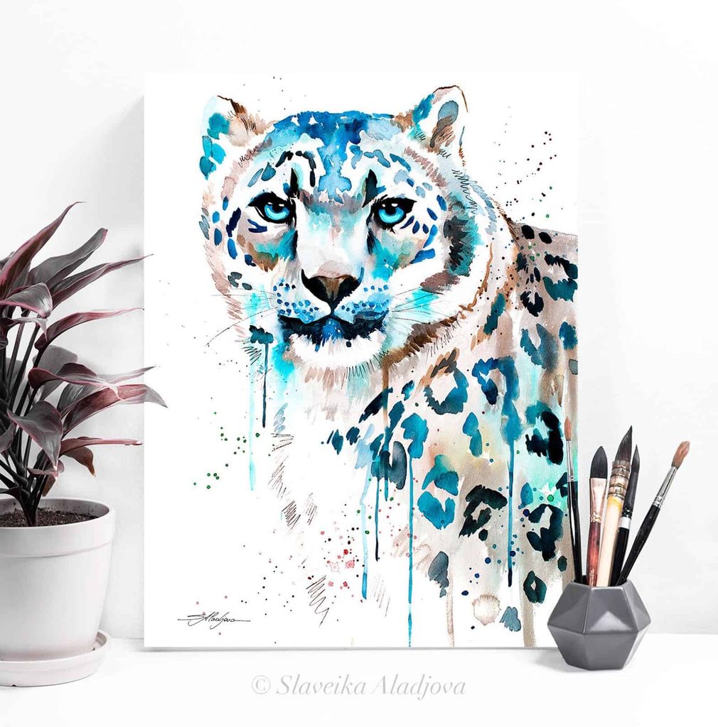 A watercolour painting of a snow leopard with blue accents is displayed upright beside a potted plant, brushes, and a pencil pot.