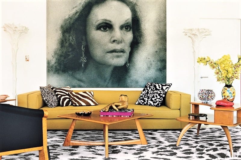 A modern living room with a yellow sofa, animal print cushions, a black-and-white rug, wooden tables, and a large greyscale portrait of a woman on the wall.
