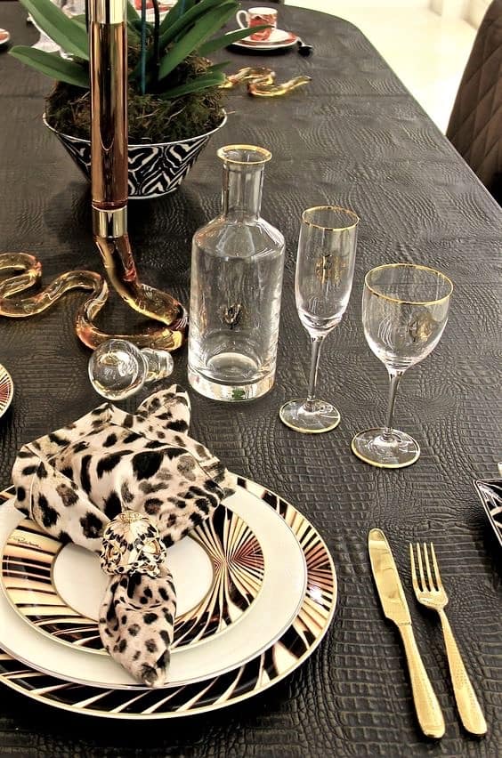 A formal dining table set with patterned plates, gold-trimmed glassware, gold cutlery, a leopard-print serviette, and a decorative snake centrepiece on a textured black tablecloth.