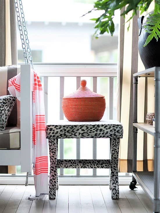 A leopard-print bench holds an orange lidded basket on a porch next to a hanging chair with a pink-striped towel and a metal shelf with a plant. Sunlight filters through curtains.