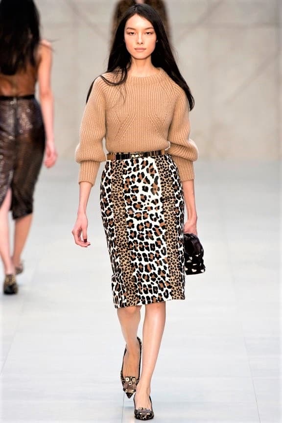 A woman walks a catwalk wearing a tan jumper, a knee-length leopard print skirt, animal print shoes, and holding a small handbag.