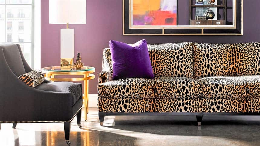 Leopard print sofa with a purple cushion, black accent chair, gold round side table, white lamp, and colourful art on a purple wall.