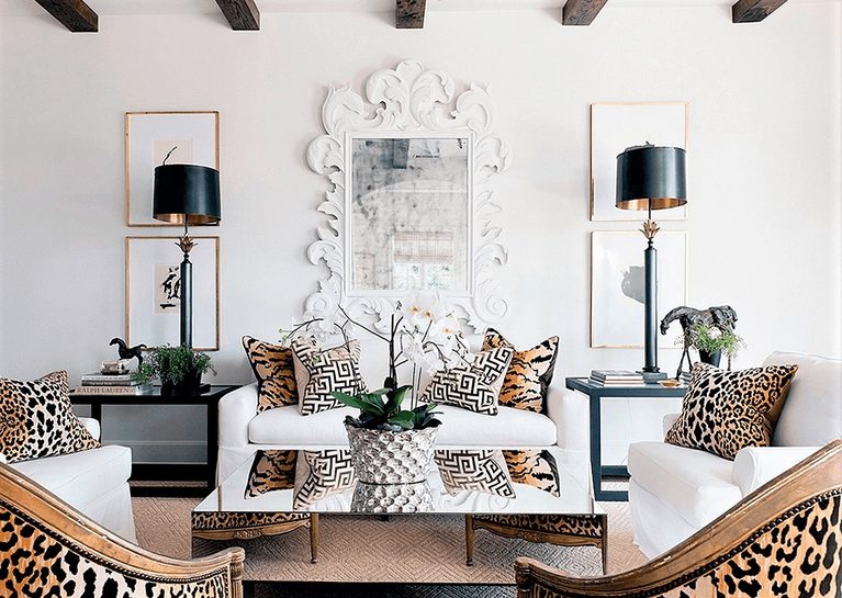 Living room with white sofas, leopard print and geometric cushions, glass coffee table, large ornate mirror, black lamps, and animal figurines on side tables.