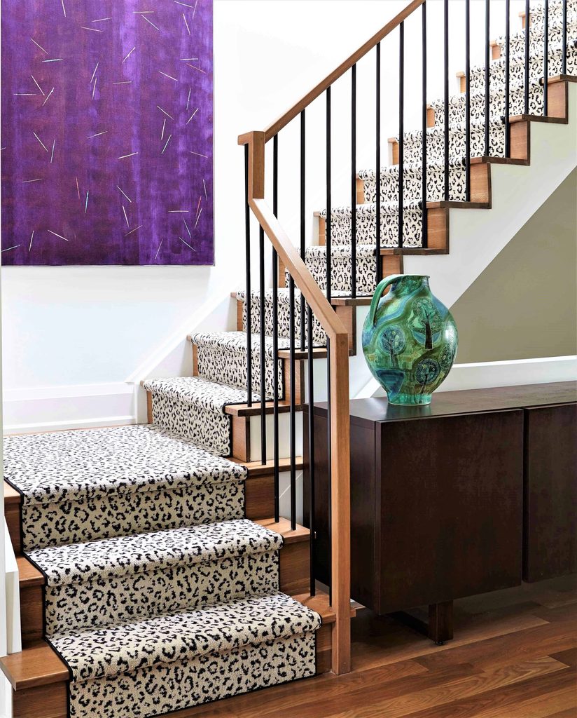 Staircase with leopard print carpet runner, black metal railing, purple abstract wall art, and a green decorative vase on a dark wood cabinet.