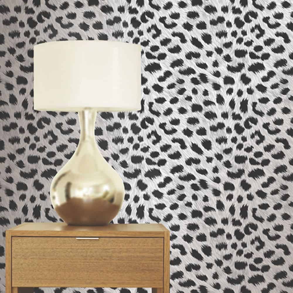 A metallic table lamp with a white shade sits on a wooden bedside table in front of a wall with a black and white spotted pattern.