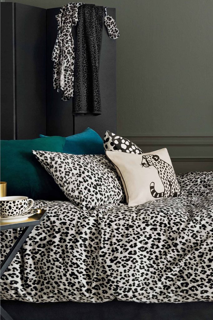 A bed with leopard print bedding, teal pillows, and a cushion with a cheetah illustration. A dressing gown and scarf with similar prints hang on a black wardrobe in the background.